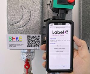 Techniker scannt mit dem Smartphone einen SHKE-Service-Sticker an einer Heizungsanlage; auf dem Display öffnet sich die Labelwin-Kontaktseite mit Auswahloptionen für Reparatur, Notdienst, Angebot und Beratung.
